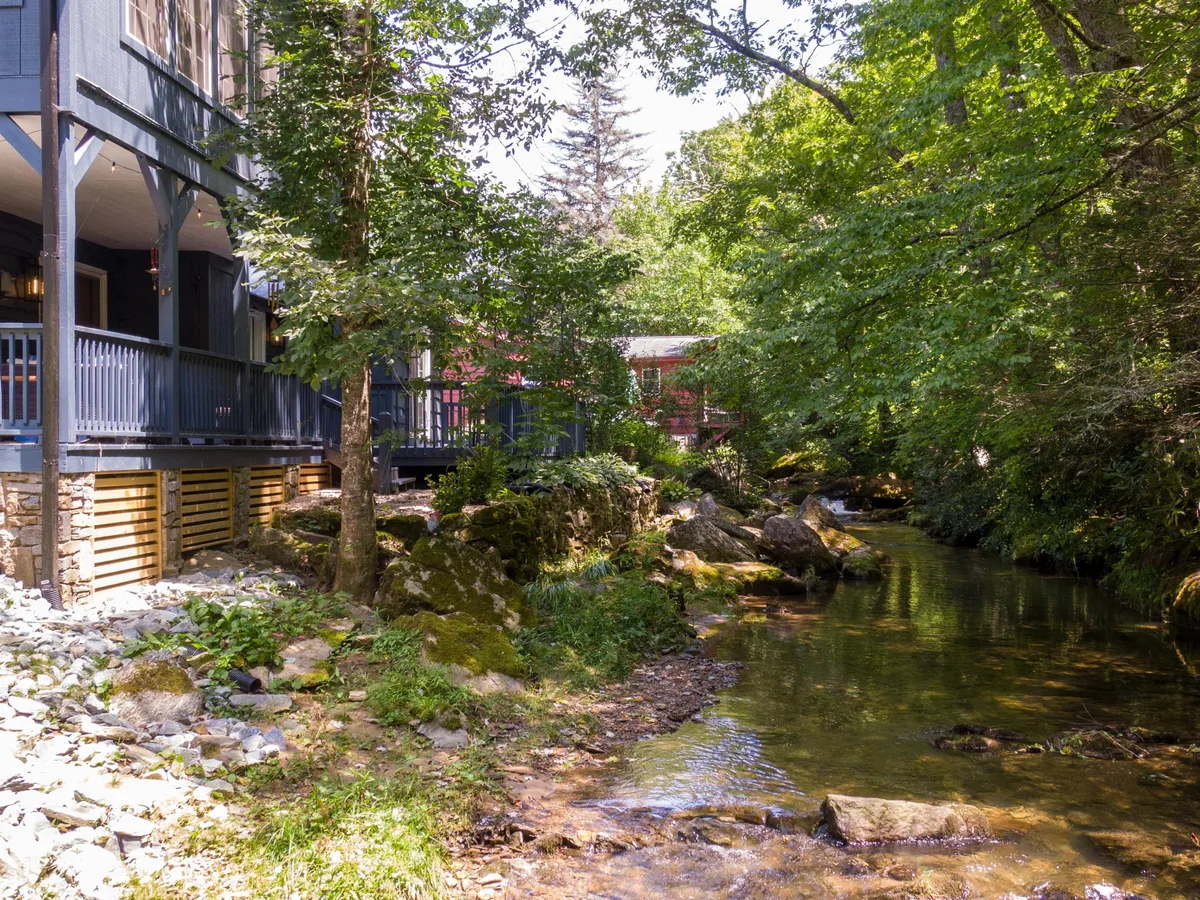 This image captures the house nestled beside a gentle stream, surrounded by mature trees and greenery. The home's proximity to the water and the open yard are clearly visible, suggesting an inviting space perfect for outdoor activities with pets.