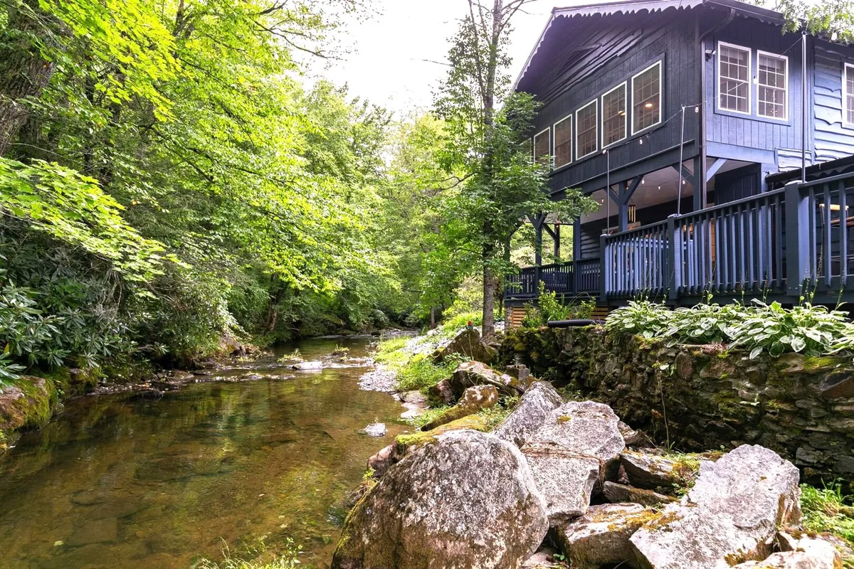 The hero image showcases a charming blue single-family house set right beside a flowing creek, surrounded by lush greenery and large natural rocks. A spacious deck looks out over the water, inviting guests to relax outdoors and take in the serene mountain views. The scene is tranquil and inviting, emphasizing the property’s direct riverfront access and beautiful natural setting.