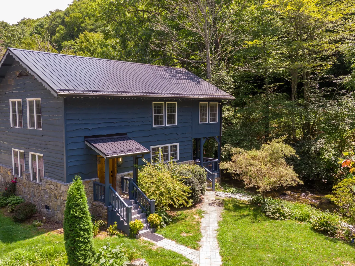 This image shows the blue wooden house with a stone foundation, surrounded by lush greenery and a welcoming pathway leading to the entrance. The porch offers an inviting spot to relax, and the setting is peaceful and perfect for group gatherings and outdoor activities.