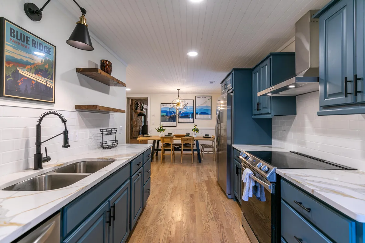 This image showcases a welcoming kitchen and dining area with marble countertops, blue cabinetry, framed artwork, and a spacious dining table set for a meal. The mix of modern appliances and stylish décor highlights a space designed for both meal preparation and communal dining.