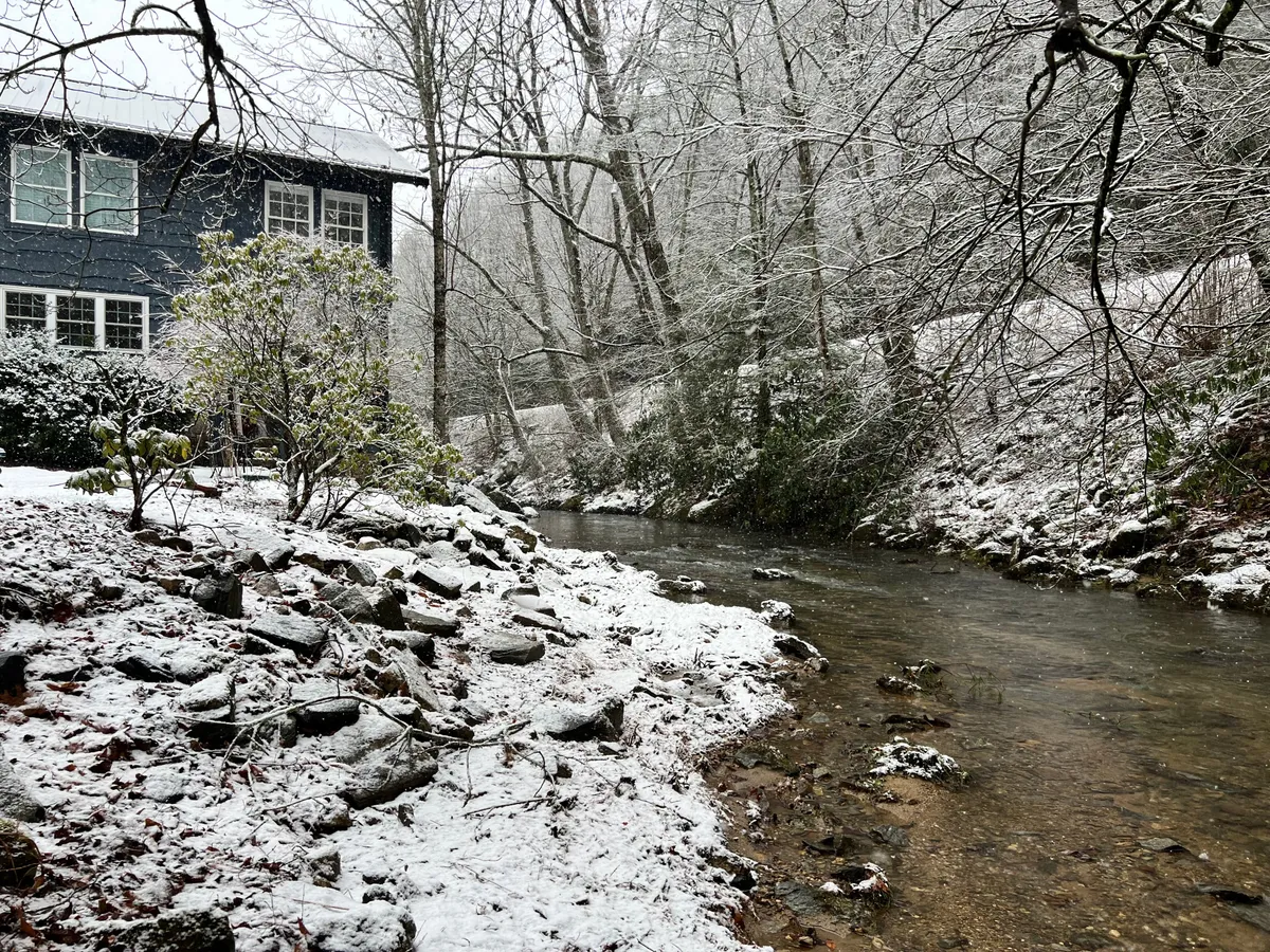 A picturesque blue house sits beside a creek, its surroundings blanketed in snow, with tall trees creating a magical winter scene. The house glows warmly against the crisp, peaceful backdrop—ideal for winter walks, cozy moments by the window, or snowy adventures.