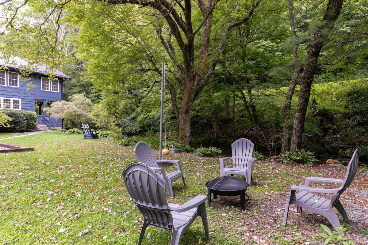 A cozy outdoor setup with Adirondack chairs encircling a fire pit, all nestled in lush greenery near a blue house. The space is inviting for gatherings, storytelling, stargazing, or simply enjoying the fresh mountain air, embodying the sense of connection to nature year-round.