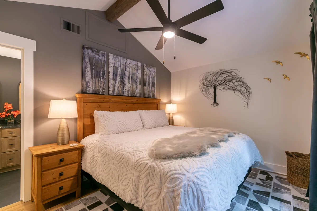 A cozy bedroom with a stylish bed, decorative pillows, soft lighting from bedside lamps, and unique artwork above the headboard. Nightstands flank the bed, offering convenience and a feeling of homely comfort.