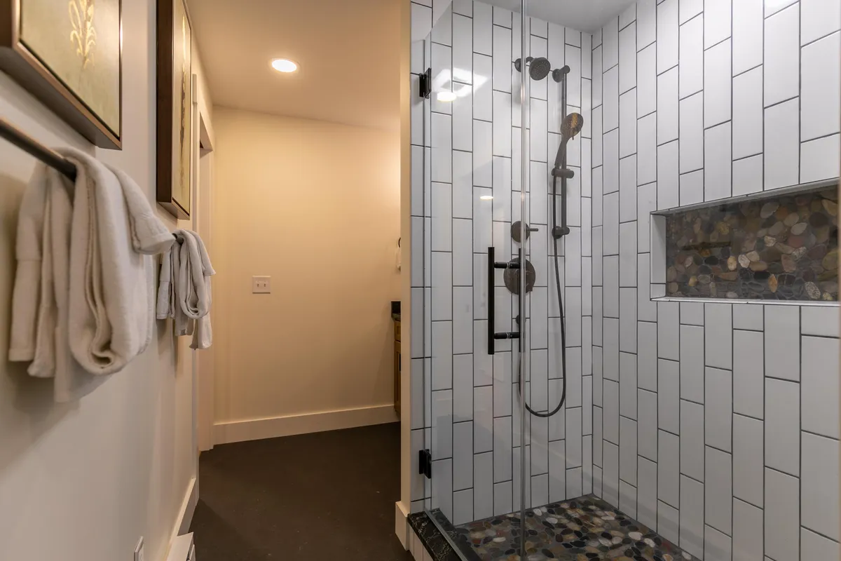 A serene bathroom with a glass shower enclosure, modern fixtures, a textured stone shower niche, and pristine white tile. The space exudes tranquility, inviting guests to unwind and refresh.