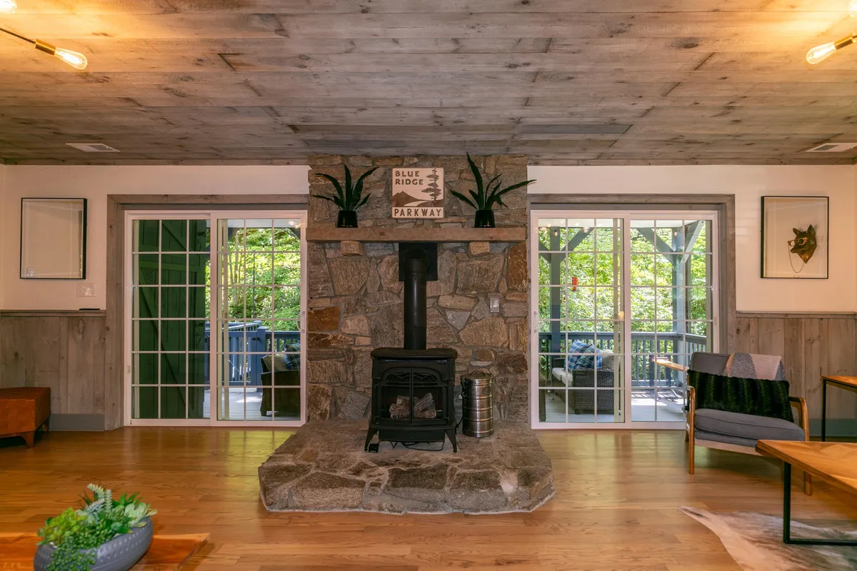 A warm and welcoming living area featuring a striking stone fireplace as the focal point. The space is adorned with wooden flooring, large windows offering views of the outdoors, and tasteful wall art. The inviting arrangement encourages gathering around the fire, reading, or entertaining guests, and the natural light enhances the room's airy, open feel.