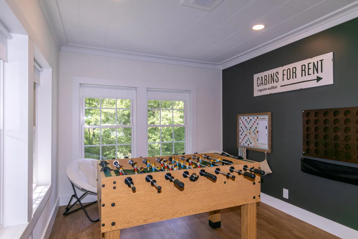 A bright sun room featuring a foosball table, views through large windows, a board game in progress, and playful decor. The room is filled with natural light and designed for energetic gatherings, making it the perfect spot for friendly competition or casual socializing with friends and family.