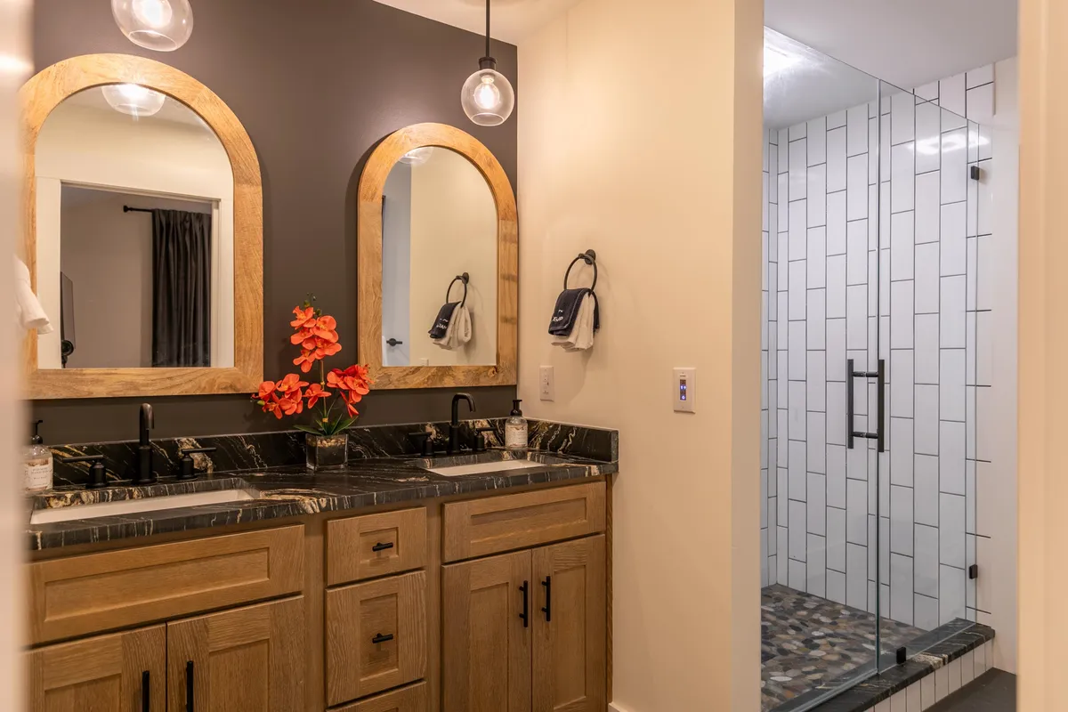 A bright, modern bathroom featuring a spacious double vanity with elegant mirrors and light fixtures. A glass shower enclosure and a decorative flower arrangement add to the calming, spa-inspired feel of the room.
